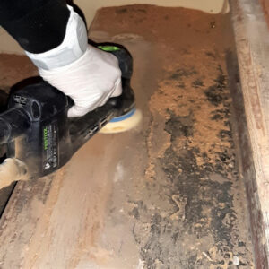 Ein Handwerker schleift alte Leimreste und lose Holzfasern von einer beschädigten Holztreppe ab, um eine saubere Basis für die Reparatur zu schaffen